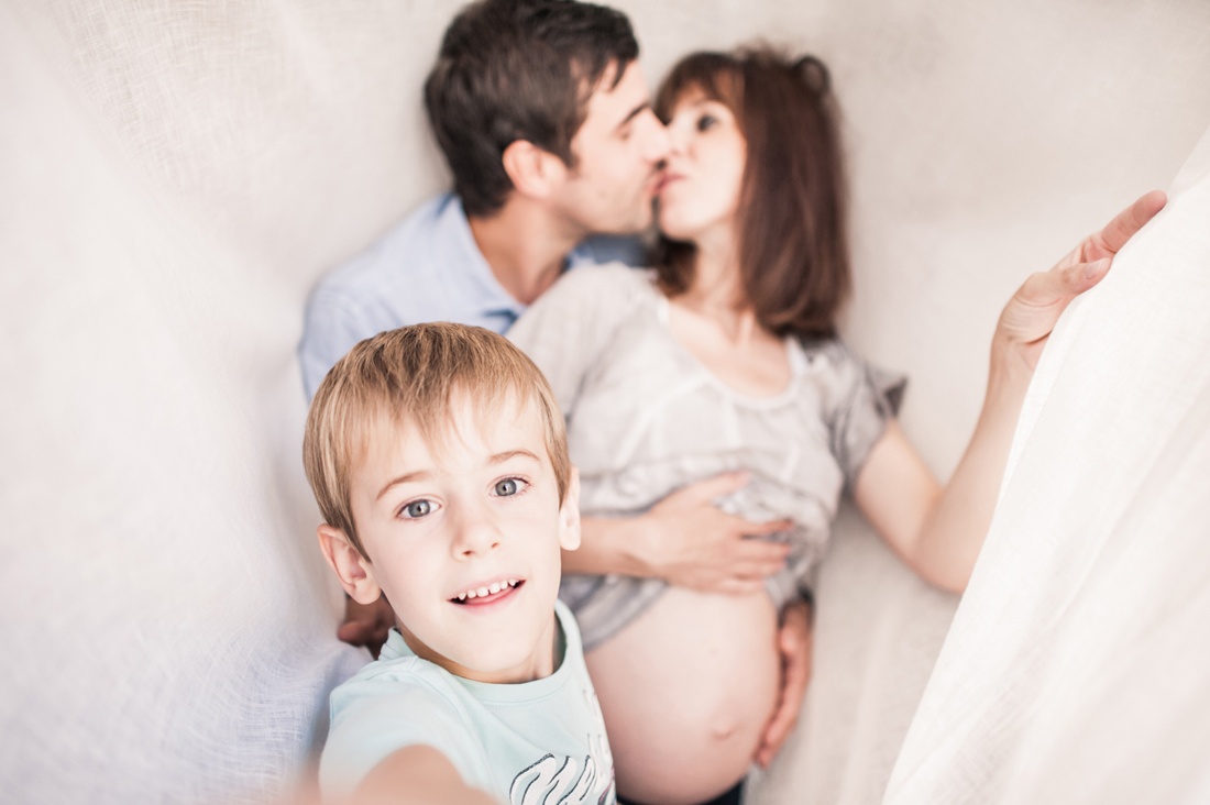 Un enfant prend une photo en selfie de lui est ses parents. La femme est enceinte. Le couple s'embrasse derrière l'enfant.