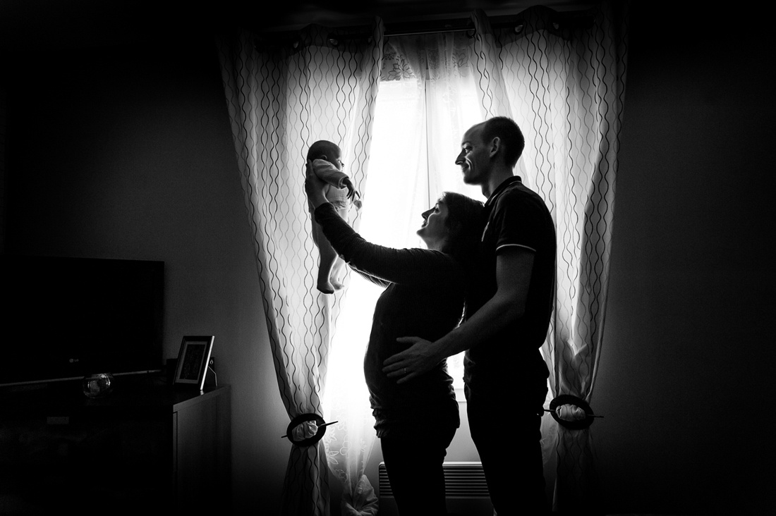 une maman et un papa devant une fenêtre en noir et blanc. Il regarde le bébé devant eux